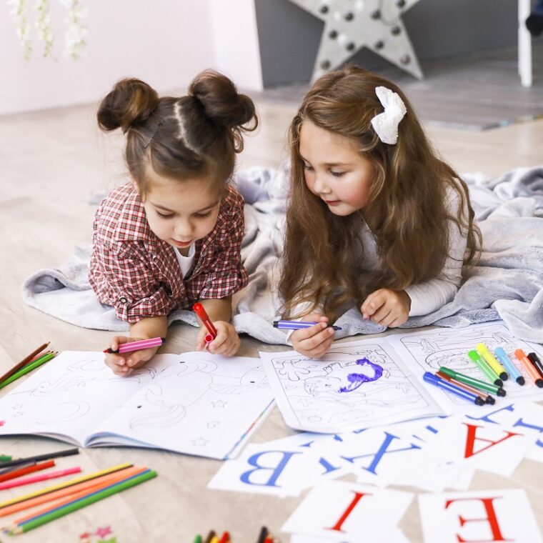 Two tiny cute girls which are drawing in the coloring book laying on the floor on the blanket and learning letters